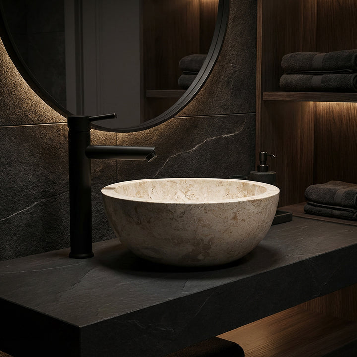 Round countertop basin made of light marble on a dark stone slab with a black fitting in an elegant bathroom ambience