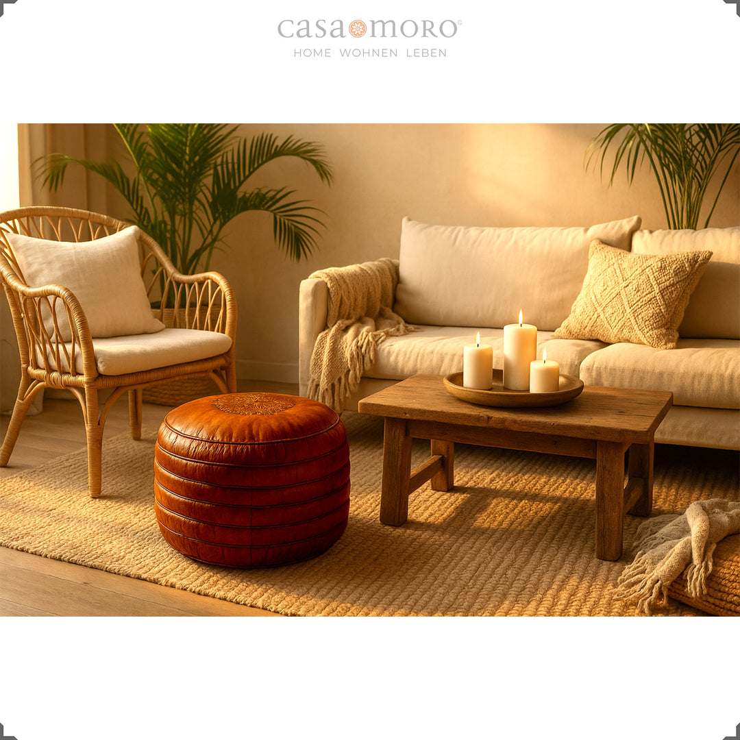 Warm living room scene with a round genuine leather pouf in brown in front of a sofa and wooden table with candles - cozy boho atmosphere in the evening light.