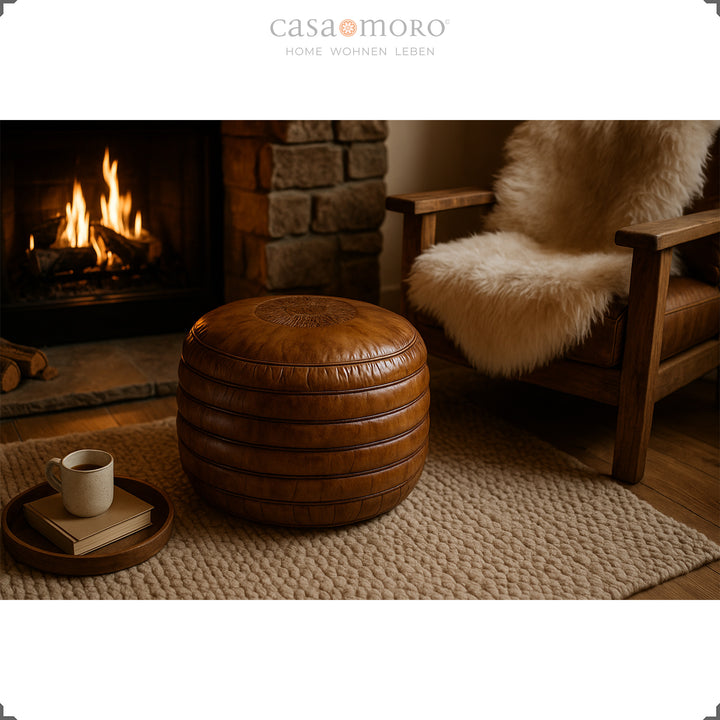 Cozy living scene with a round leather pouf in warm brown in front of the fireplace, next to it a wooden armchair with a fur cushion and a cup on a tray - atmospheric ambience.