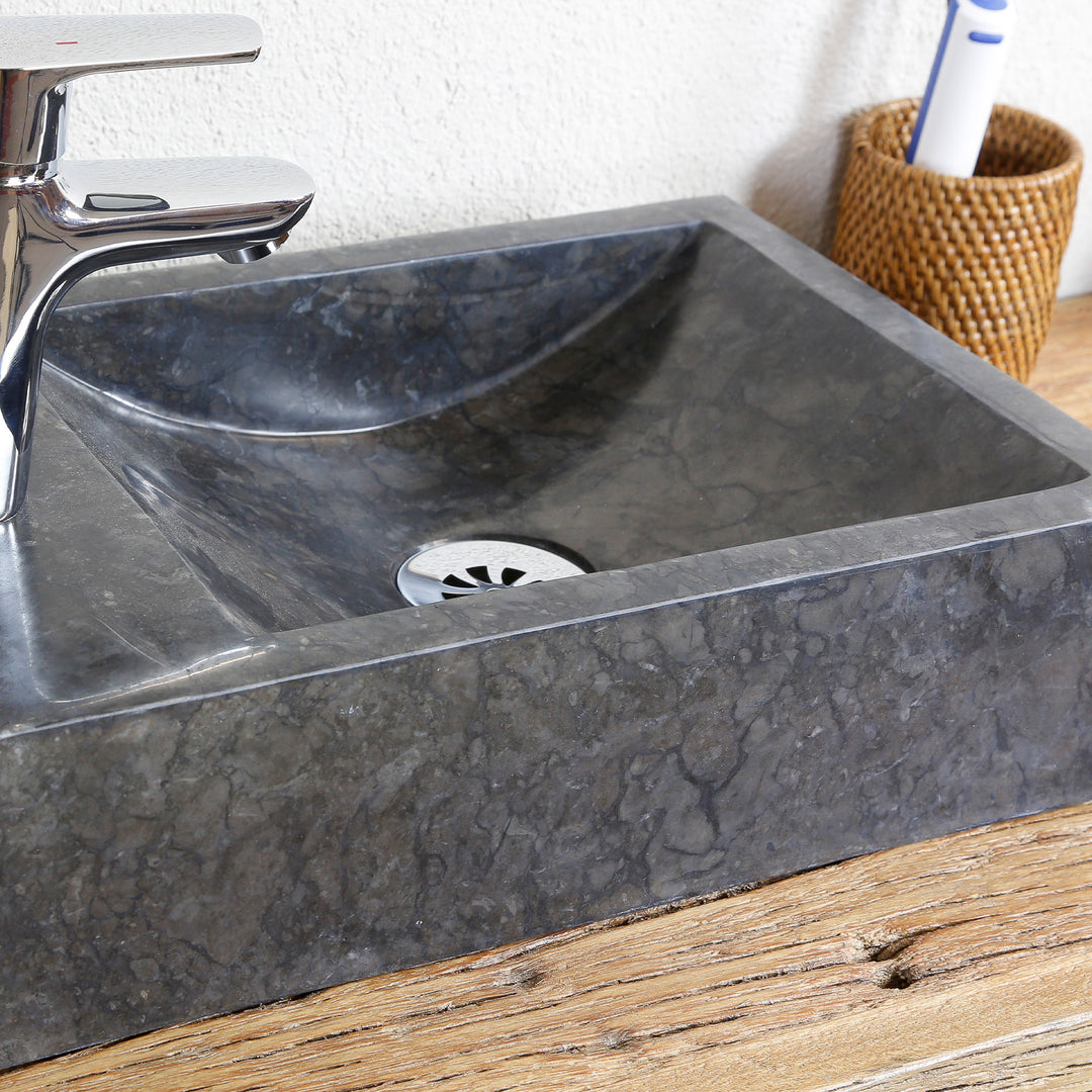 Angular natural stone countertop basin with dark grain in a detailed view, with stainless steel drain and chrome-plated fitting on a wooden plate