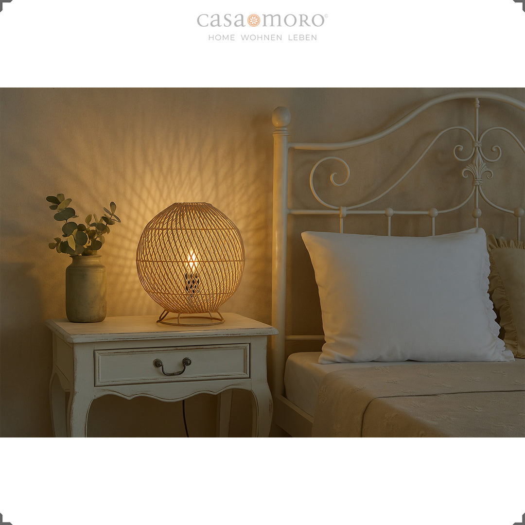 Round rattan bedside lamp on white wooden table next to bed, warm light and soft atmosphere