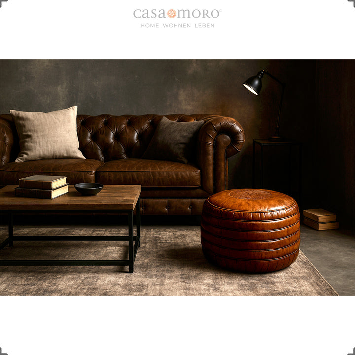 Elegant living room with Chesterfield sofa, coffee table and round leather pouf in brown - stylish scene in industrial and modern-rustic style.