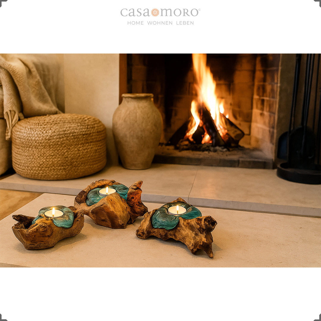 Three tea light holders made of glass and root wood in front of a burning fireplace, their soft light underlines the cozy living atmosphere.