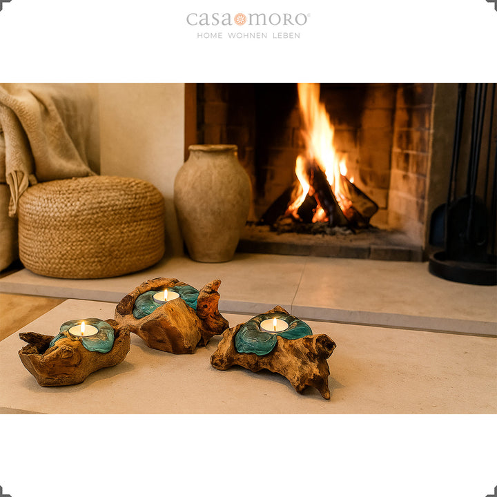 Three tea light holders made of glass and root wood in front of a burning fireplace, their soft light underlines the cozy living atmosphere.