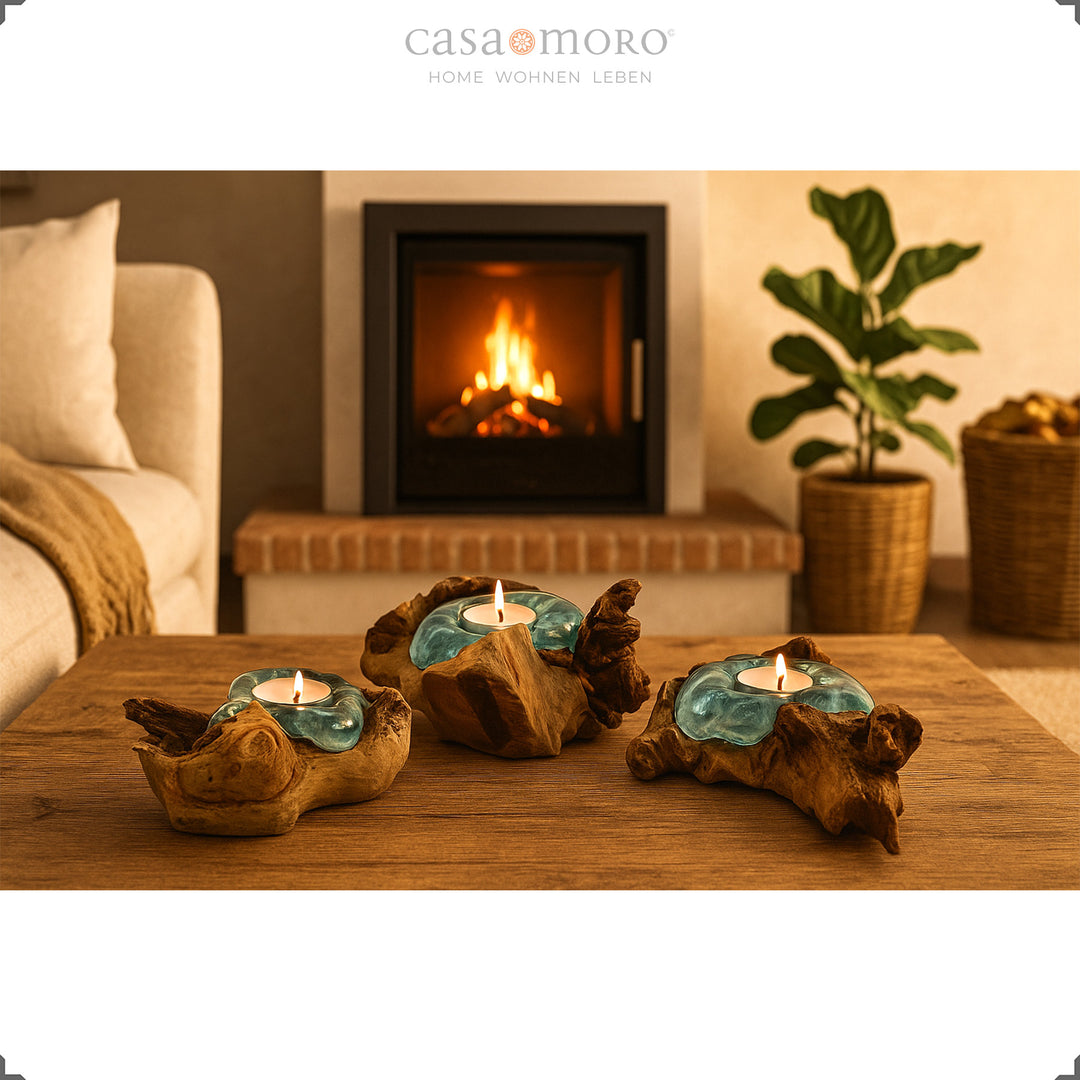 Three tea light holders made of glass and root wood on a wooden table, their warm light complements the cozy fireplace atmosphere in the living room.