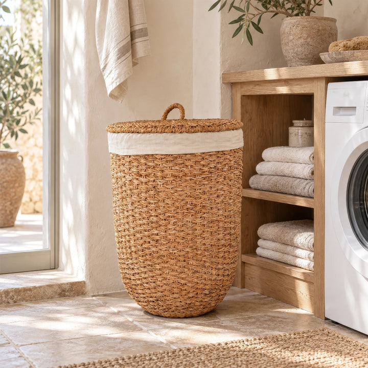 A laundry basket with a lid made of natural-colored seagrass next to a towel rack and a washing machine in the laundry room.